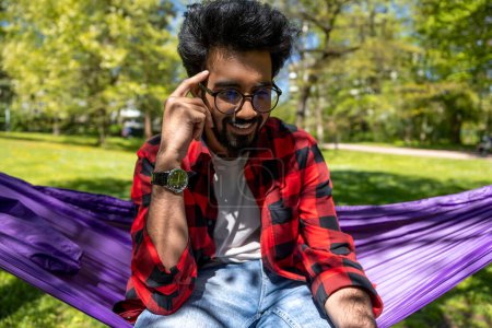 Foto de Joven hombre hindú en camisa a cuadros se siente relajado en hamaca en el parque - Imagen libre de derechos