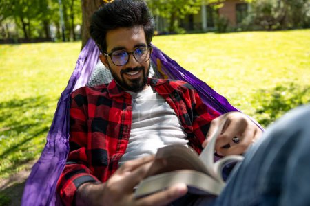 Téléchargez les photos : Jeune homme aux cheveux foncés lisant un livre tout en restant allongé dans un hamac - en image libre de droit