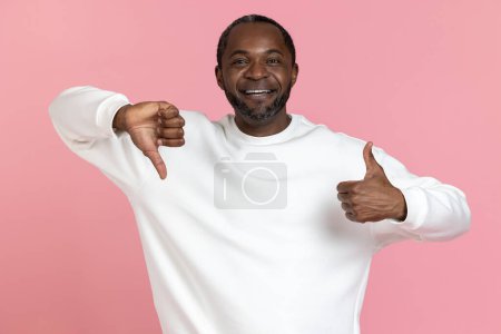 Foto de Hombre negro sonriente con sudadera blanca mostrando como y gesto de aversión aislado sobre fondo rosa. - Imagen libre de derechos
