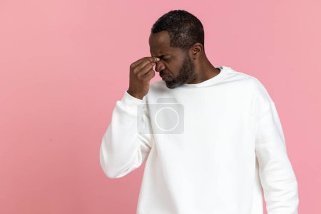 Foto de Triste hombre negro cansado usando sudadera blanca con la cabeza dolorosa aislada sobre fondo rosa. - Imagen libre de derechos