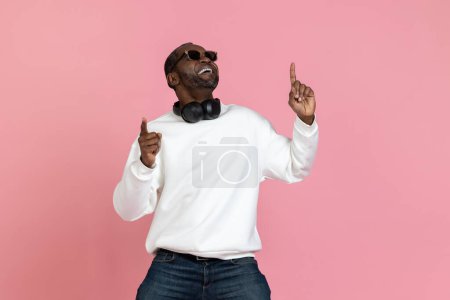 Foto de Hombre negro sonriente con sudadera blanca apuntando hacia el espacio de copia aislado sobre fondo rosa. - Imagen libre de derechos