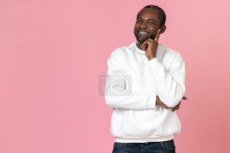 Foto de Hombre negro sonriente con sudadera blanca sosteniendo barbilla aislada sobre fondo rosa. - Imagen libre de derechos