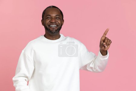Foto de Hombre negro alegre con sudadera blanca apuntando hacia el espacio de copia aislado sobre fondo rosa. - Imagen libre de derechos
