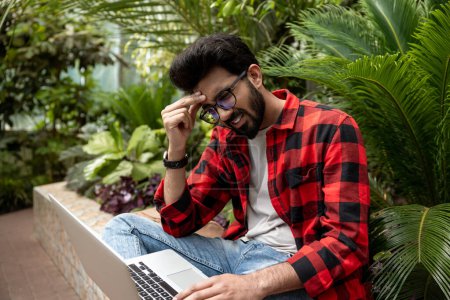 Téléchargez les photos : Homme en chemise à carreaux rouges travaillant sur ordinateur portable et l'air fatigué - en image libre de droit