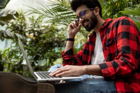 Téléchargez les photos : Homme en chemise à carreaux rouges travaillant sur ordinateur portable dans un jardin - en image libre de droit