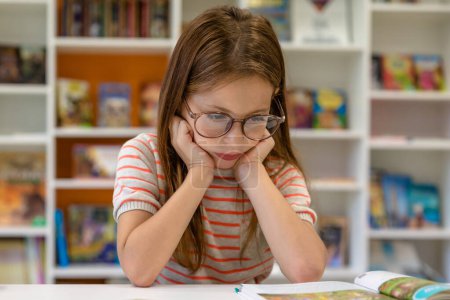 Foto de Una chica aburrida leyendo un libro en la biblioteca de la escuela. Creatividad e imaginación infantil. - Imagen libre de derechos