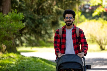 Téléchargez les photos : Jeune père. Jeune homme en chemise à carreaux portant un landau dans le parc - en image libre de droit