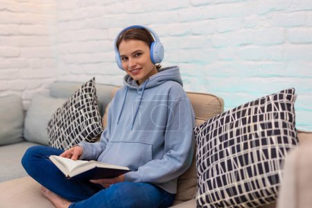Téléchargez les photos : Jeune femme écoutant la musique des écouteurs et du livre de lecture tout en étant assis sur le canapé dans le salon. - en image libre de droit