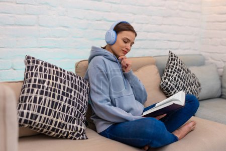 Photo for Woman listening music in headphones and reading book while sitting on couch in home interior. - Royalty Free Image