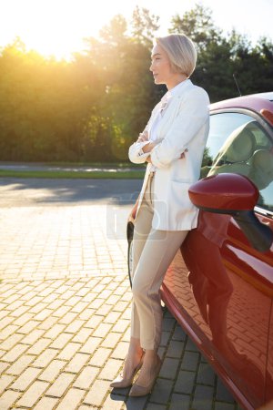 Téléchargez les photos : Femme d'affaires blonde portant un costume de style officiel blanc près de son automobile rouge sur le parking - en image libre de droit