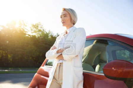 Téléchargez les photos : Belle femme adulte debout avec les bras croisés près de sa nouvelle voiture rouge - en image libre de droit