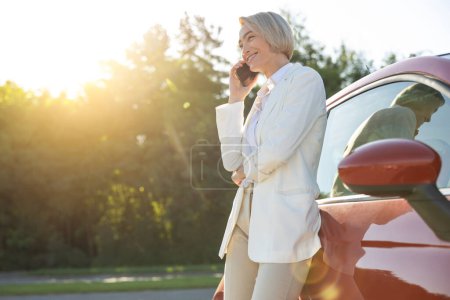 Foto de Impresionante mujer hablando en el teléfono móvil mientras está de pie en el aparcamiento cerca de su coche rojo - Imagen libre de derechos