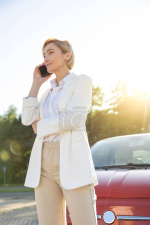 Foto de Impresionante mujer hablando en el teléfono móvil mientras está de pie en el aparcamiento cerca de su coche rojo - Imagen libre de derechos