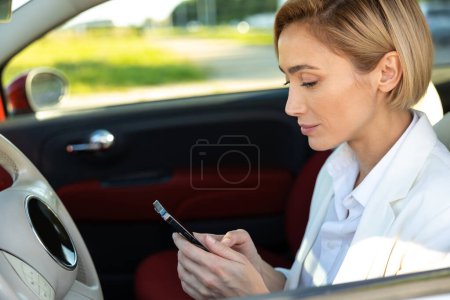 Téléchargez les photos : Vue latérale de la femme blonde utilisant un smartphone pendant un conseil d'affaires en automobile - en image libre de droit