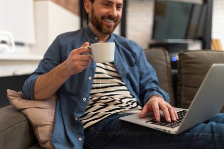 Foto de Joven barbudo guapo hombre sentado en casa y trabajando en el ordenador portátil - Imagen libre de derechos