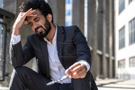 Photo for Dark-haired young man in suit sitting on steps outside and suffering from headache - Royalty Free Image
