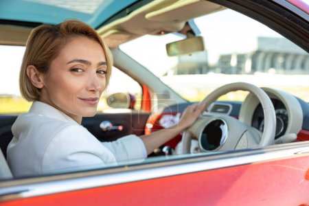 Foto de Atractiva mujer sonriente con traje de estilo oficial manteniendo el volante en el coche - Imagen libre de derechos