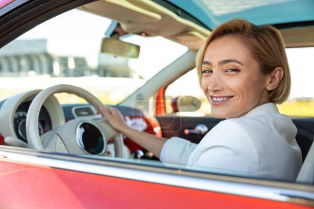Téléchargez les photos : Femme d'affaires souriante conduisant une voiture assise sur le siège du conducteur dans sa voiture rouge voyageant seule - en image libre de droit