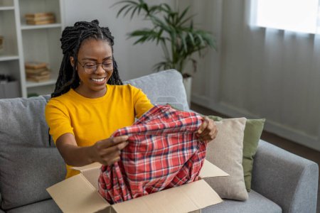 Photo for Smiling dark-skinned woman opening box with clothes and looking contented - Royalty Free Image
