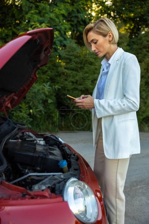 Photo for Blonde young woman holding phone searching emergency car service in internet while standing near broken car with open hood - Royalty Free Image