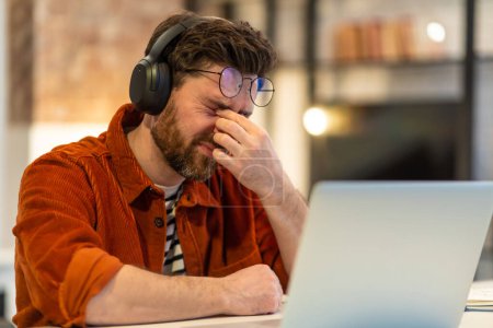 Téléchargez les photos : Homme barbu assis à l'ordinateur portable et regardant fatigué après le travail - en image libre de droit