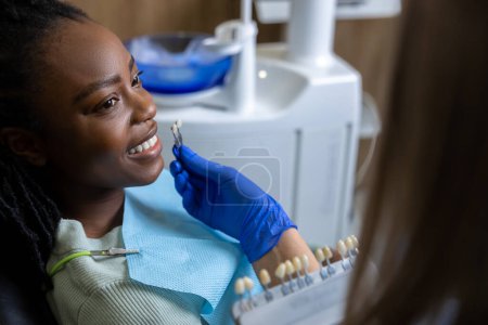 Téléchargez les photos : Dentiste méconnaissable tenant le tableau de guide d'ombrage dentaire pour le patient féminin pendant le traitement de blanchiment dans la clinique moderne - en image libre de droit