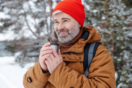 Foto de Un día frío. Hombre barbudo de pelo gris con sombrero rojo y termos en las manos en un bosque de invierno - Imagen libre de derechos