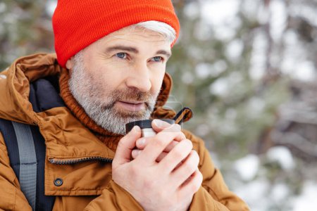 Foto de Tomando té caliente. Hombre barbudo de pelo gris con sombrero rojo y termos en las manos en un bosque de invierno - Imagen libre de derechos