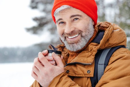 Téléchargez les photos : Du thé chaud. Homme barbu aux cheveux gris en chapeau rouge avec thermos dans les mains dans une forêt d'hiver - en image libre de droit
