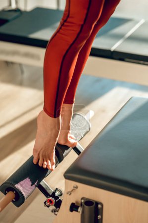 Téléchargez les photos : Femme méconnaissable en legging rouge fait des exercices pour renforcer les muscles des jambes sur Pilates réformateur en studio de sport - en image libre de droit