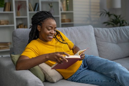 Téléchargez les photos : Jeune femme mignonne à la peau foncée lisant un livre et souriant - en image libre de droit