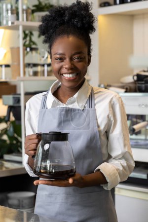 Téléchargez les photos : Serveur femme multiculturelle gai tenant pot en verre avec café brassage debout près comptoir café - en image libre de droit