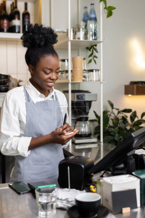 Téléchargez les photos : Femme multiraciale barman dans un café portant tablier ordre d'écriture dans le carnet de service pour le client - en image libre de droit