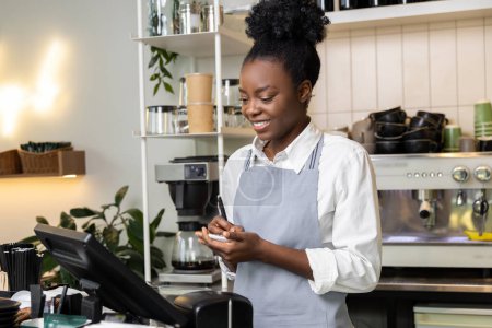 Téléchargez les photos : Femme multiraciale barman dans un café portant tablier ordre d'écriture dans le carnet de service pour le client - en image libre de droit