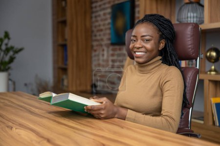 Téléchargez les photos : Femme gaie avec dreadlocks assis à la table lecture livre pigiste s'éduquer sur les stratégies d'affaires - en image libre de droit