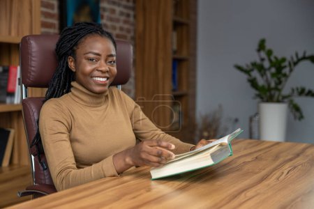 Téléchargez les photos : Femme joyeuse avec des dreadlocks assis à la table lecture étudiant livre approfondissant ses connaissances à des fins éducatives - en image libre de droit