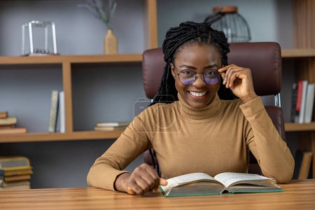 Téléchargez les photos : Femme ravie avec dreadlocks assis à la table lecture de livre chercheur de connaissances constamment s'éduquer dans son lieu de travail - en image libre de droit