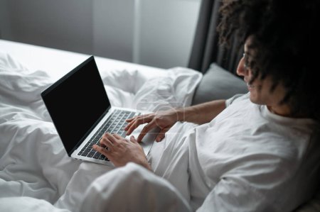 Photo for Remote work. Brunette man in white tshirt lying in bed and working on laptop - Royalty Free Image