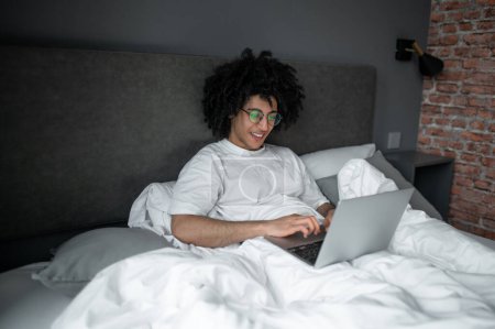 Trabajo remoto. Hombre morena en camiseta blanca acostado en la cama y trabajando en el ordenador portátil