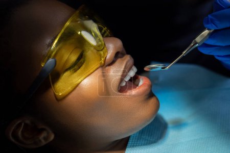 Foto de Paciente femenina multicultural brillantemente iluminada abre la boca mientras está sentada en la silla del dentista en la clínica - Imagen libre de derechos
