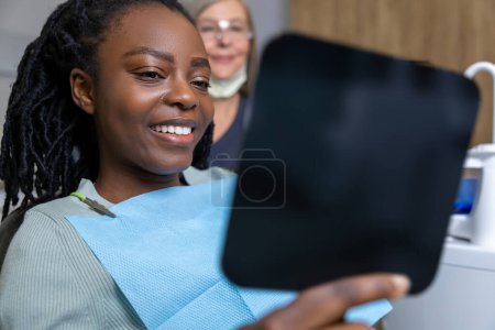 Téléchargez les photos : Femme multiraciale joyeuse assis dans la chaise dans le cabinet de stomatologie regardant miroir à son sourire parfait - en image libre de droit