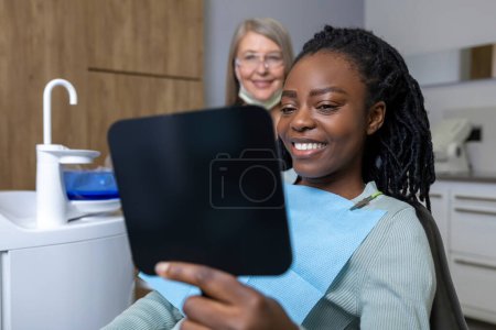 Téléchargez les photos : Heureuse patiente multiculturelle regardant miroir après un traitement dentaire en clinique - en image libre de droit