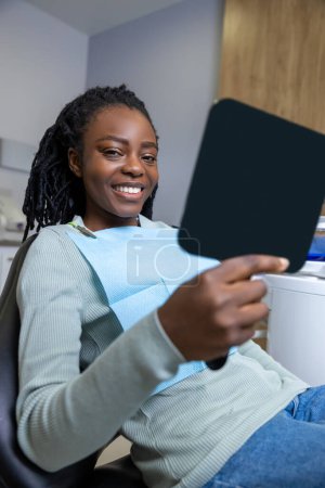 Téléchargez les photos : Femme multiraciale joyeuse assis dans la chaise dans le cabinet de stomatologie regardant miroir à son sourire parfait - en image libre de droit