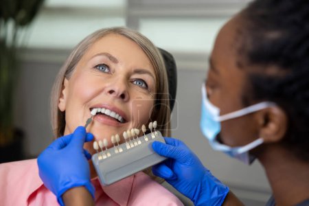 Téléchargez les photos : Patiente souriante dans le siège des dentistes tandis que le médecin orthodontiste choisissant la nuance de placage dentaire selon le nuancier - en image libre de droit