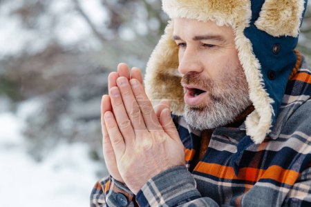 Cold. Mature man in warm hat warming his hands with his breath