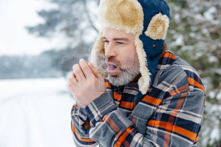 Cold. Mature man in warm hat warming his hands with his breath