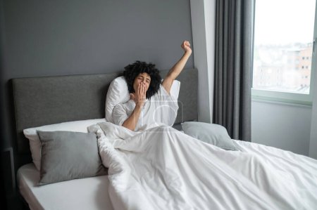 Waking up. Curly-haired brunette young man waking up in a hotel bedroom