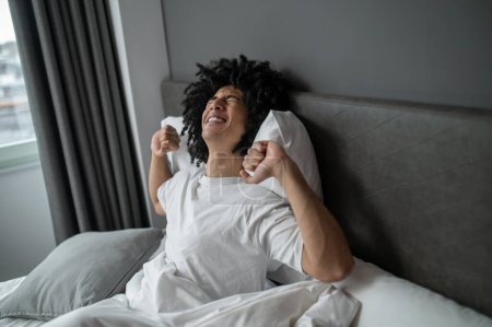 Waking up. Curly-haired brunette young man waking up in a hotel bedroom
