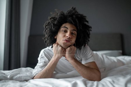 Slow mode. Young curly-haired man lying in bed and looking relaxed and sleepy