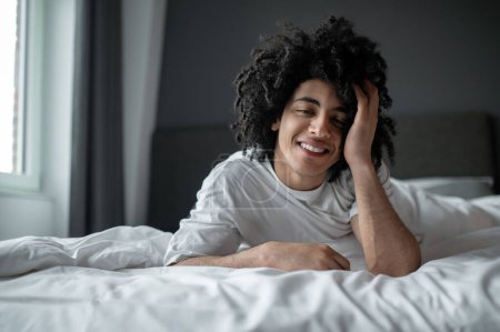 Slow mode. Young curly-haired man lying in bed and looking relaxed and sleepy